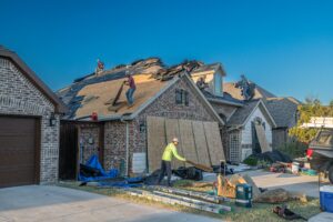Workers are removing old shingles and repairing the roof of a brick house. Some workers are on the roof, while one is on the ground collecting debris. Construction materials and tools are scattered around the driveway.
