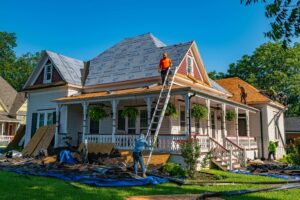 Workers replace the roof of a large, light-colored house with a wraparound porch; some are on ladders, while others are on the roof, surrounded by roofing materials and debris on the ground.