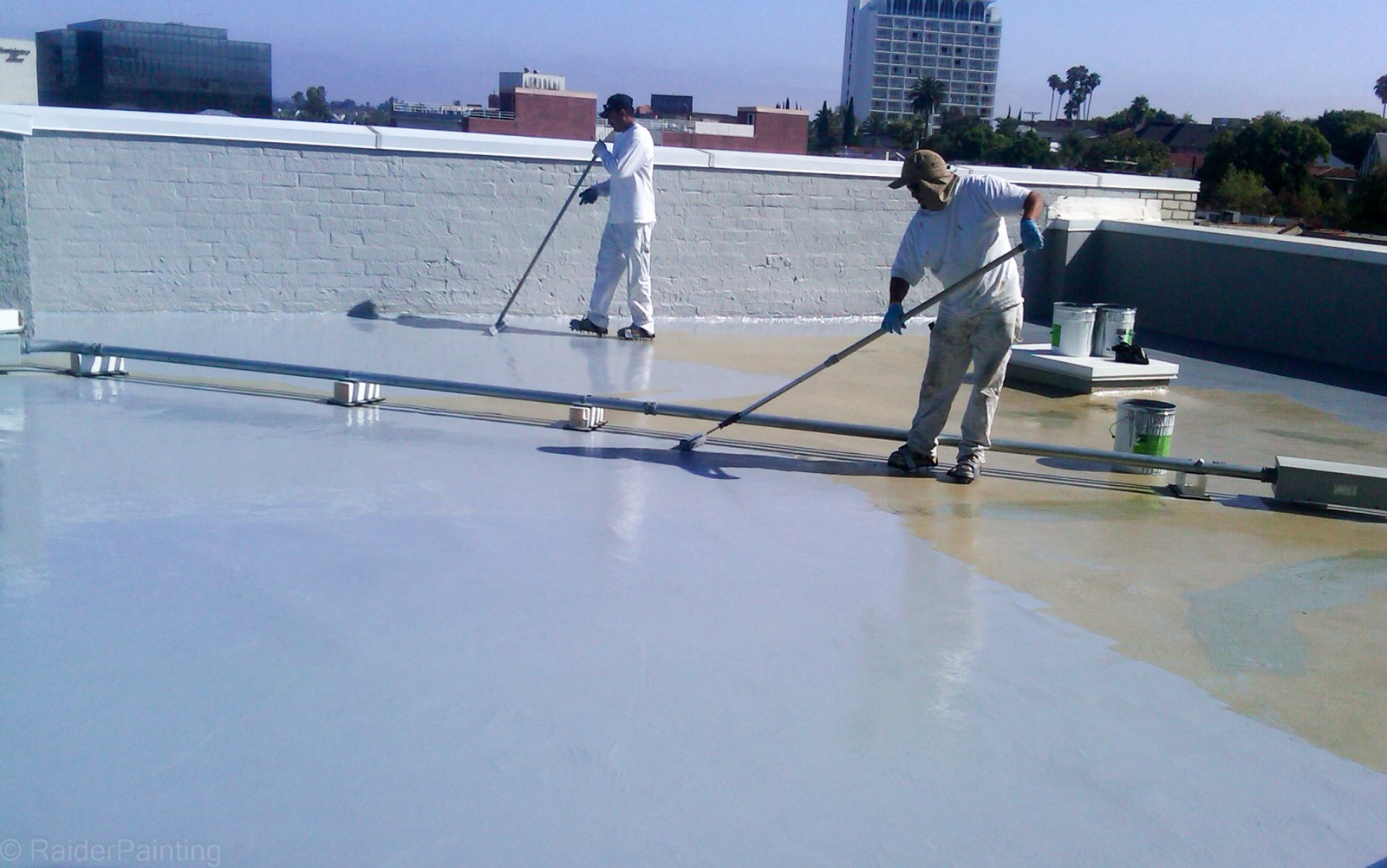 Two workers wearing hats and white clothing apply a waterproof coating to a flat rooftop using long rollers on handles. Paint cans and tools are nearby, and buildings are visible in the background under a clear sky.